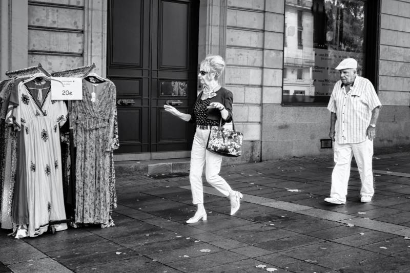 Un homme et une femme âgés et élégants marchent sur le trottoir d'une rue piétonne en pierre, passant devant la devanture d'un magasin et un portant de robes de braderie affichant un prix de 20 euros. La femme au premier plan, portant des lunettes de soleil, un collier de perles et un sac à fleurs, esquisse un geste de la main en regardant vers les vêtements, tandis que l'homme à sa suite, en chemise à rayures et casquette blanche, la regarde marcher. Photographié en noir et blanc très contrasté.